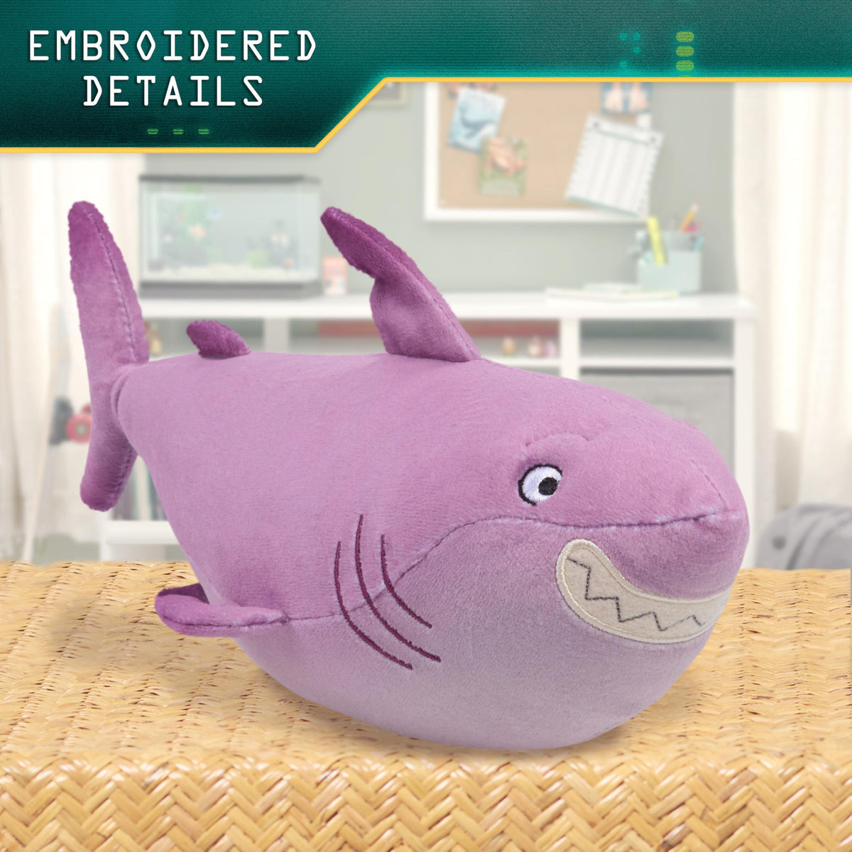Plush purple shark toy with embroidered eyes, mouth, and gill details on a woven surface with a room background.